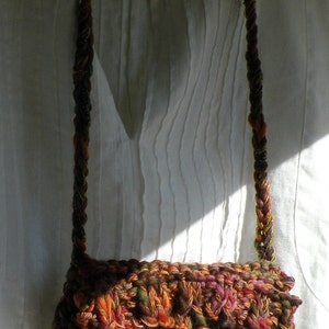 May include: A brown, green, orange, and red crocheted bag with a brown button. The bag is hanging from a long, brown, and orange crocheted strap.