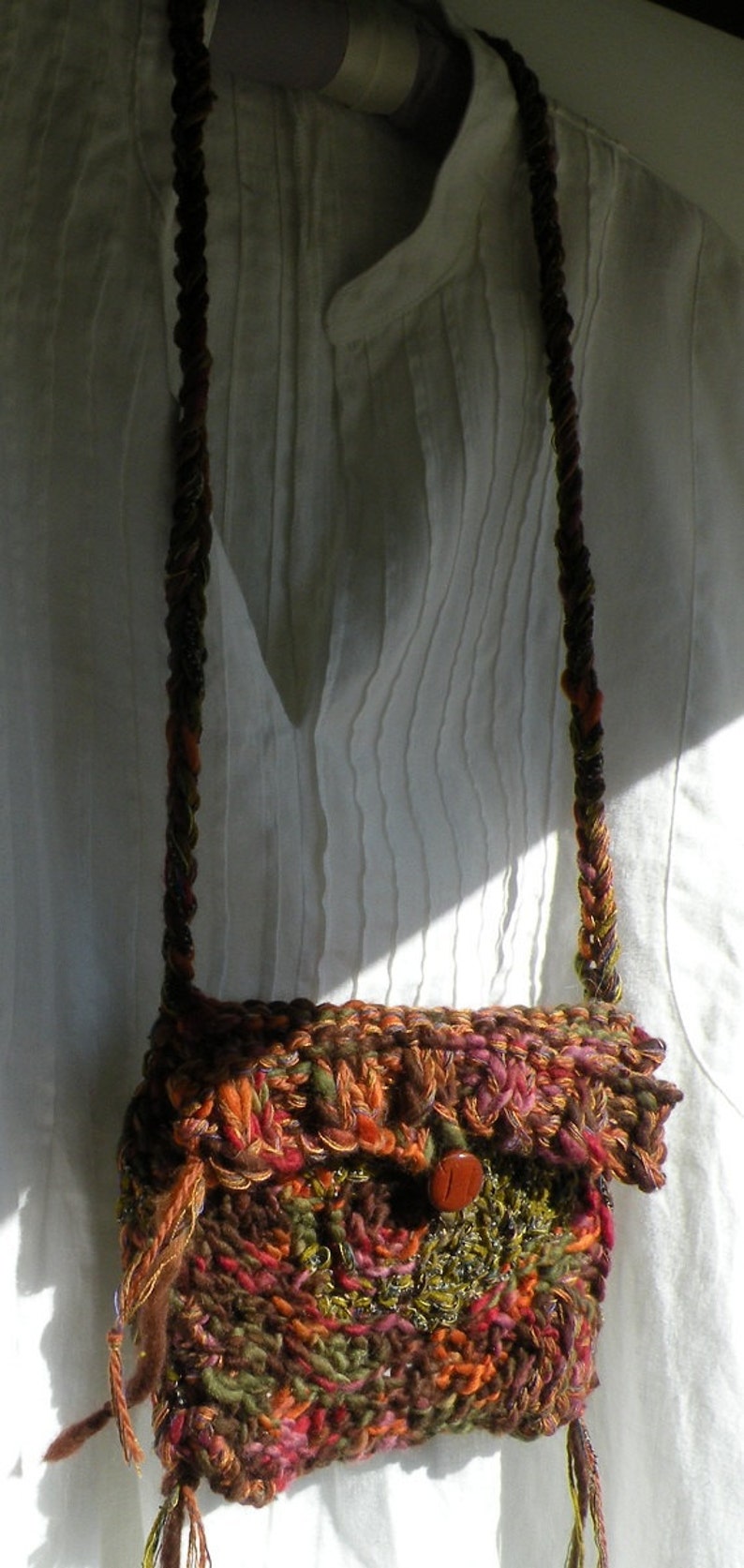 May include: A brown, green, orange, and red crocheted bag with a brown button. The bag is hanging from a long, brown, and orange crocheted strap.