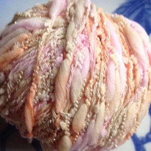 Puede incluir: Un ovillo de hilo en una mezcla de colores rosa, melocot&oacute;n y crema. El hilo tiene un aspecto esponjoso y texturizado y est&aacute; decorado con peque&ntilde;as cuentas brillantes.