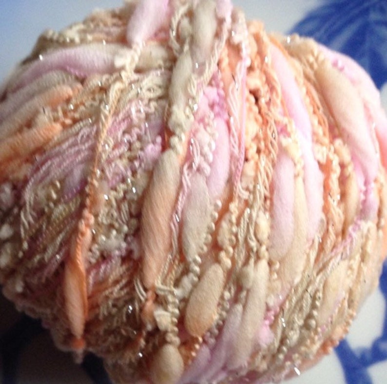 Puede incluir: Un ovillo de hilo en una mezcla de colores rosa, melocot&oacute;n y crema. El hilo tiene un aspecto esponjoso y texturizado y est&aacute; decorado con peque&ntilde;as cuentas brillantes.