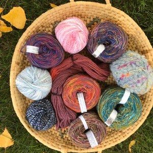 May include: A basket filled with colorful yarn balls in various textures, including a fuzzy blue ball, a striped blue and gold ball, and a pink ball. The yarn is arranged in a circular pattern with a brown scarf in the center.