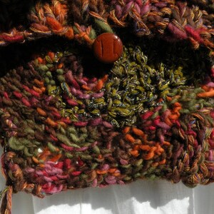May include: A close-up of a knitted bag with a brown, green, and red color scheme. The bag has a button closure and a fringe detail.
