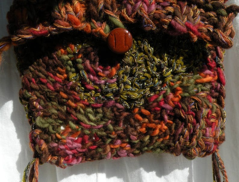 May include: A close-up of a knitted bag with a brown, green, and red color scheme. The bag has a button closure and a fringe detail.
