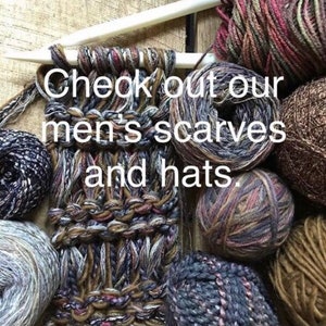 Puede incluir: Primer plano de una bufanda tejida con diferentes tonos de hilo marr&oacute;n, gris y blanco. La bufanda est&aacute; rodeada de ovillos de lana de diferentes colores y texturas. El texto "Check out our men's scarves and hats." es visible en la imagen.