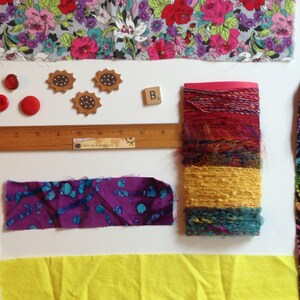 May include: A collection of craft supplies including floral fabric, red buttons, wooden embellishments, a wooden ruler, a scrabble tile, and colorful yarn. Various fabric scraps in purple, yellow, and patterned designs are also present.