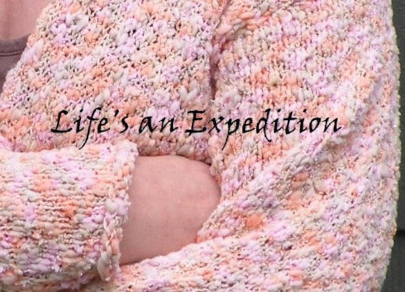 Puede incluir: Primer plano de un c&aacute;rdigan de punto rosa y melocot&oacute;n con un aspecto texturizado y esponjoso. El c&aacute;rdigan est&aacute; doblado y el texto "Life's an Expedition" es visible en la tela.