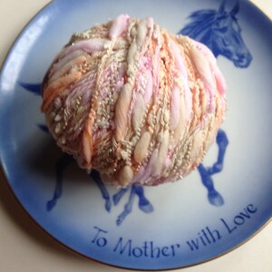 Puede incluir: Una bola de hilo rosa, melocot&oacute;n y blanco con una textura brillante, sentada en un plato de porcelana azul y blanco con las palabras "To Mother with Love" escritas en &eacute;l.
