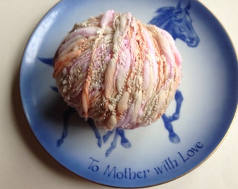 May include: A ball of pink, peach, and white yarn with a sparkly texture, sitting on a blue and white porcelain plate with the words "To Mother with Love" written on it.