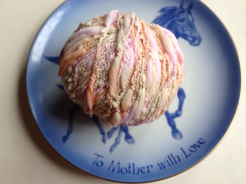 Puede incluir: Una bola de hilo rosa, melocot&oacute;n y blanco con una textura brillante, sentada en un plato de porcelana azul y blanco con las palabras "To Mother with Love" escritas en &eacute;l.