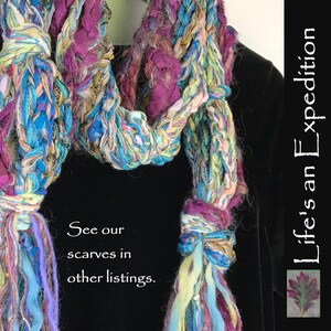 May include: A multi-colored crocheted scarf with long fringe tassels. The scarf is made with a variety of yarns, including blue, green, purple, and white. The scarf is draped over a black garment. The text "See our scarves in other listings." is printed below the scarf. The text "Life's an Expedition" is printed on the right side of the image.
