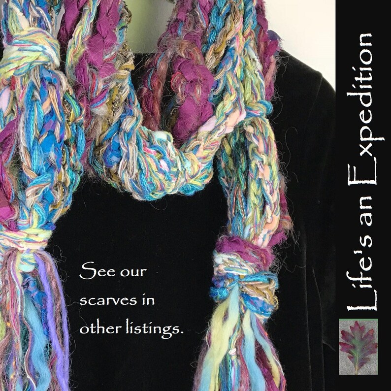 May include: A multi-colored crocheted scarf with long fringe tassels. The scarf is made with a variety of yarns, including blue, green, purple, and white. The scarf is draped over a black garment. The text "See our scarves in other listings." is printed below the scarf. The text "Life's an Expedition" is printed on the right side of the image.