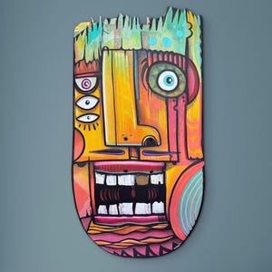 May include: A colorful, abstract wooden mask with a stylized face. The mask features a large open mouth with white teeth, multiple eyes, and a prominent nose. The color palette includes yellow, orange, pink, and blue, with a rough, textured top edge.