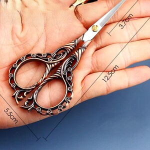 May include: Antique-style scissors with ornate, floral-patterned handles. The blades are silver and the handles are copper-toned. The scissors are 5.5 inches long and 3.7 inches wide.