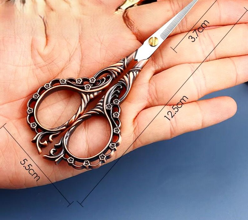 May include: Antique-style scissors with ornate, floral-patterned handles. The blades are silver and the handles are copper-toned. The scissors are 5.5 inches long and 3.7 inches wide.