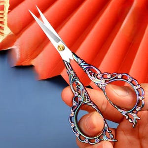 May include: A pair of small, ornate scissors with a rainbow iridescent finish. The handles are decorated with a floral design and rhinestones.