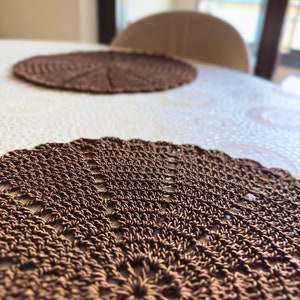May include: Two brown, crocheted placemats on a white tablecloth. The placemats have a circular shape with a detailed, openwork design. The focus is on the intricate texture and the warm brown color of the placemats, suggesting a handmade craft.