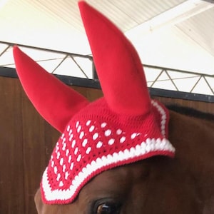 Op de afbeelding: Een rood en wit gehaakt paardenvliegengordijn met oorbedekkingen. De kap heeft een rode basis met witte stippen en een witte rand. De oren van het paard zijn bedekt met rode stof. Het paard is bruin.