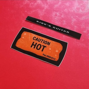 Könnte beinhalten: Zwei schwarze Etiketten auf rotem Untergrund. Auf einem Etikett steht "KIRK'S GUITAR" in weißer Schrift. Das andere Etikett hat einen orangefarbenen Hintergrund mit den Worten "CAUTION HOT" und der Teilenummer "#850-0113" in schwarzer Schrift.