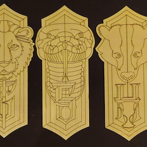 Puede incluir: Cuatro marcapáginas de madera con diseños grabados. Cada uno presenta un animal diferente: un león, una serpiente, un tejón y un águila. Los marcapáginas también incluyen letras y símbolos estilizados.