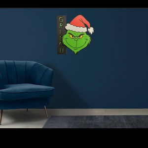 May include: A green Grinch face wearing a red Santa hat, with the word "GRINCH" vertically on a dark brown background. The Grinch is mounted on a dark blue wall. A blue chair and a dark grey rug are in the foreground.