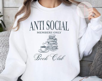 Puede incluir: Sudadera blanca con el texto "ANTI SOCIAL MEMBERS ONLY Book Club" en azul oscuro. El diseño incluye una pila de libros y una taza de té. La parte superior se usa con vaqueros azules.