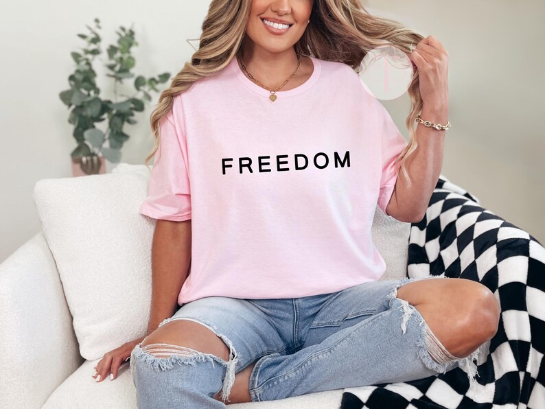 Peut inclure: Un t-shirt rose clair avec le mot "FREEDOM" imprim&eacute; en noir. Le t-shirt est port&eacute; avec un jean bleu. La personne est assise sur un canap&eacute; blanc avec une couverture &agrave; carreaux noirs et blancs.