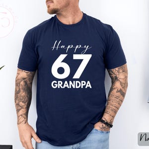May include: Navy blue t-shirt with the text "Happy 67 Grandpa" in white script and block letters. The shirt is a solid color and has a crew neck. The image also shows a plant and a denim jacket.