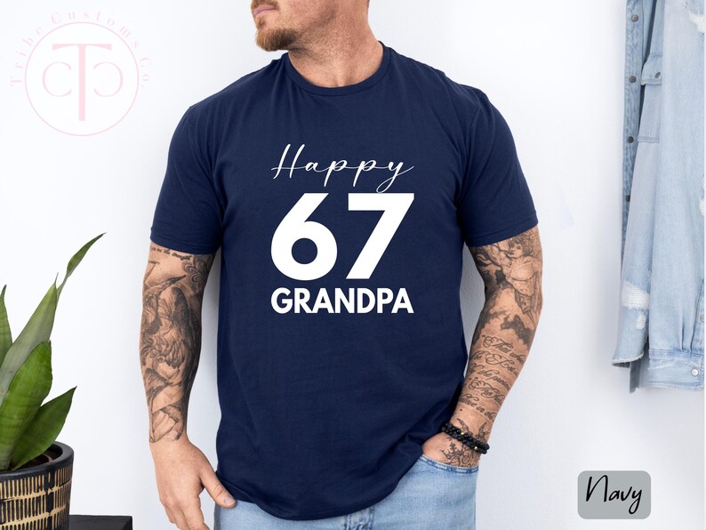 May include: Navy blue t-shirt with the text "Happy 67 Grandpa" in white script and block letters. The shirt is a solid color and has a crew neck. The image also shows a plant and a denim jacket.