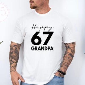 May include: White t-shirt with the text "Happy 67 Grandpa" in black. The number 67 is large and bold, with "Happy" above and "Grandpa" below in a smaller font. The shirt is being worn by a person.