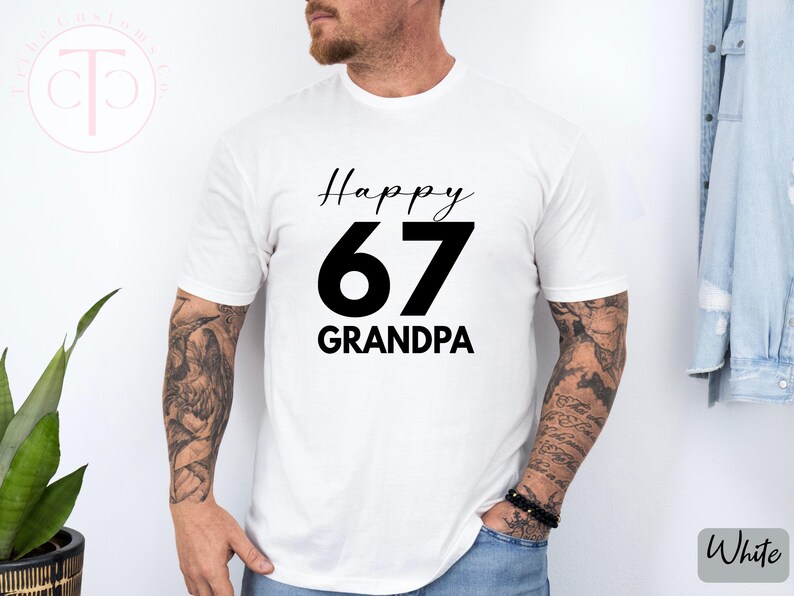 May include: White t-shirt with the text "Happy 67 Grandpa" in black. The number 67 is large and bold, with "Happy" above and "Grandpa" below in a smaller font. The shirt is being worn by a person.