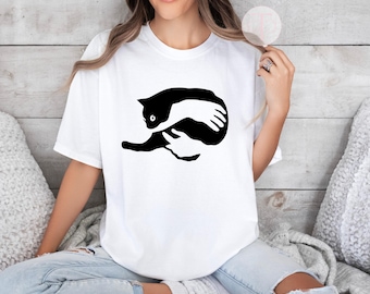 May include: A white t-shirt with a black and white graphic of a cat being held by a hand. The cat is curled up and the hand is wrapped around it.