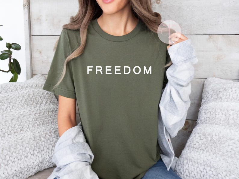Peut inclure: T-shirt vert olive avec le mot "FREEDOM" imprim&eacute; en blanc. Le t-shirt est de style col rond. Le mod&egrave;le porte une veste grise et blanche claire sur le t-shirt.