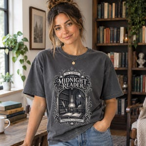 Könnte beinhalten: Ein anthrazitfarbenes Kurzarm-T-Shirt mit einem detaillierten Grafikdruck einer Bibliotheksszene und dem Text "EX LIBRIS THE MIDNIGHT READER PRIVATE LIBRARY". Das Design zeigt ein aufgeschlagenes Buch, eine Lampe, einen Globus.