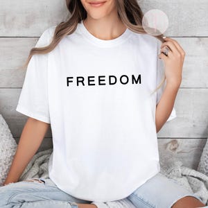 Peut inclure: T-shirt blanc avec le mot &laquo; FREEDOM &raquo; imprim&eacute; en lettres noires. Le t-shirt est de style col rond et semble &ecirc;tre fait d'une mati&egrave;re douce. La personne porte un jean bleu.