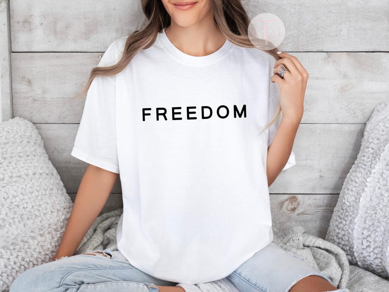 Peut inclure: T-shirt blanc avec le mot &laquo; FREEDOM &raquo; imprim&eacute; en lettres noires. Le t-shirt est de style col rond et semble &ecirc;tre fait d'une mati&egrave;re douce. La personne porte un jean bleu.