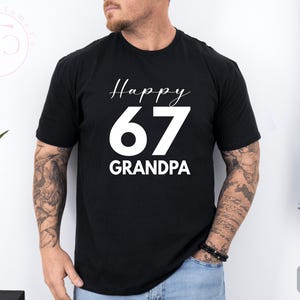 May include: Black t-shirt with the text "Happy 67 Grandpa" in white script and bold font. The number 67 is large and centered. The shirt is short-sleeved and appears to be made of cotton.