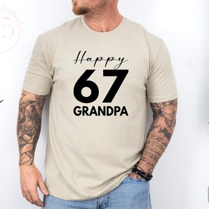 May include: Sand-colored t-shirt with the text "Happy 67 GRANDPA" in black. The number 67 is large and bold. The shirt is short-sleeved and appears to be made of a soft material. The model is wearing a black bracelet.