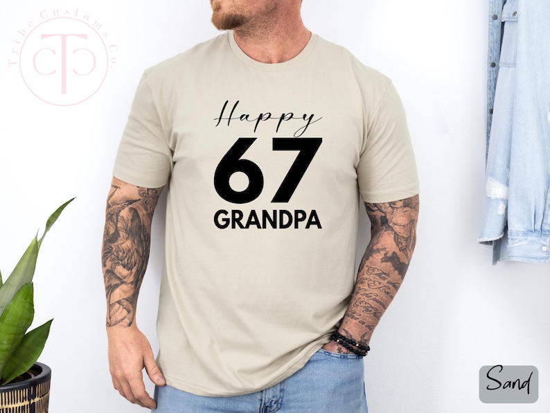 May include: Sand-colored t-shirt with the text "Happy 67 GRANDPA" in black. The number 67 is large and bold. The shirt is short-sleeved and appears to be made of a soft material. The model is wearing a black bracelet.
