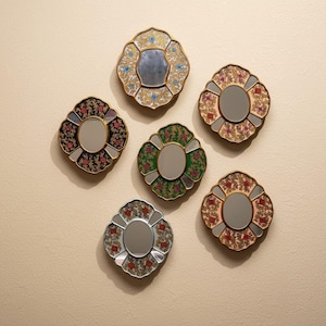 Pode incluir: Seis espelhos decorativos de parede com desenhos florais. Os espelhos têm uma borda ondulada e um espelho central circular. A paleta de cores inclui dourado, vermelho, verde, preto e prateado. Os espelhos estão dispostos em um padrão circular.