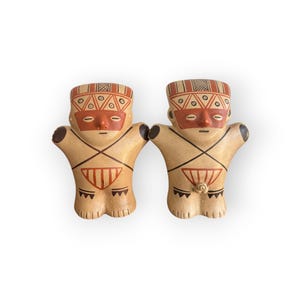 Könnte beinhalten: Zwei keramische Figuren in Beige mit roten und schwarzen Akzenten. Die Figuren haben eine stilisierte menschliche Form mit ausgestreckten Armen. Sie weisen geometrische Muster auf Kopf und Körper sowie rote Gesichtsmarkierungen auf.
