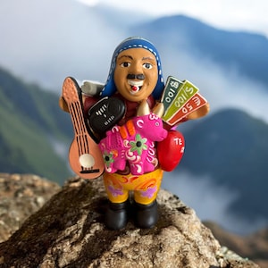 May include: A colorful ceramic figurine of a man holding a guitar, money, and a pink donkey. The man wears a blue hat, red shirt, and yellow pants. The donkey is decorated with floral patterns. The figurine has a cheerful expression.