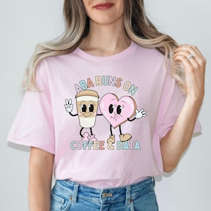 Puede incluir: Camiseta rosa con un gráfico de una taza de café y una rosquilla en forma de corazón con el texto "ABA runs on coffee & data".