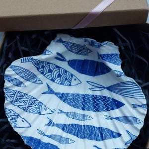 May include: A white ceramic seashell dish with a blue and white fish pattern. The dish is in a brown box with black shredded paper.