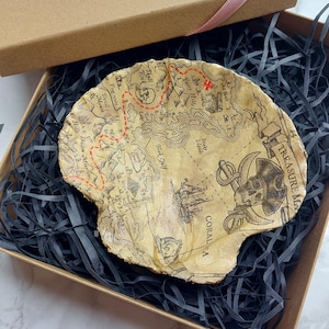 May include: A treasure map-shaped dish made of resin, with a brown and beige colour scheme. The dish features a pirate skull and crossbones design, along with the words "TREASURE MAP" and "CORALA".