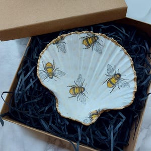 May include: A white seashell dish with a gold rim and a bee design. The dish is in a brown box with black shredded paper.