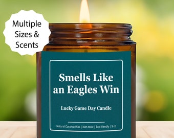 Può includere: Un barattolo di candela verde acqua con un coperchio marrone e un'etichetta bianca che recita "Smells Like an Eagles Win" e "Lucky Game Day Candle". L'etichetta riporta anche "Natural Coconut Wax | Non-toxic | Eco-friendly | 9 oz".