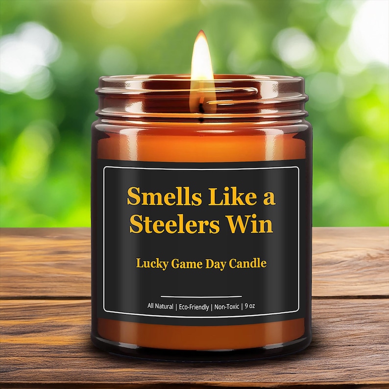 Wonderscape Wicks candle shop on Etsy presenting lit 9oz amber jar soy wax candle with text smells like a Steelers win lucky game day candle on a wood table in a garden. Pittsburgh Football NFL sports gift for husband wife fiance boyfriend