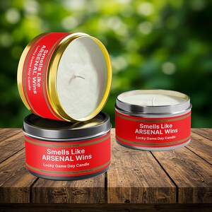 Wonderscape Wicks candle shop on Etsy presenting 8 oz tin candles in black, gold, and silver colors with text smells like Arsenal Wins Lucky Game Day Candle on a table. The all natural cotton-wick candle can be personalized with customer's custom text. The design is aimed at Premier League Football Soccer fans with Arsenal red and white colors. Gift for wife boyfriend husband girlfriend brother sister Bukayo Saka Martin Odegaard Declan Rice Gabriel Martinelli Thierry Henry Dennis Bergkamp