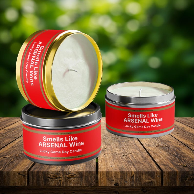 Wonderscape Wicks candle shop on Etsy presenting 8 oz tin candles in black, gold, and silver colors with text smells like Arsenal Wins Lucky Game Day Candle on a table. The all natural cotton-wick candle can be personalized with customer's custom text. The design is aimed at Premier League Football Soccer fans with Arsenal red and white colors. Gift for wife boyfriend husband girlfriend brother sister Bukayo Saka Martin Odegaard Declan Rice Gabriel Martinelli Thierry Henry Dennis Bergkamp