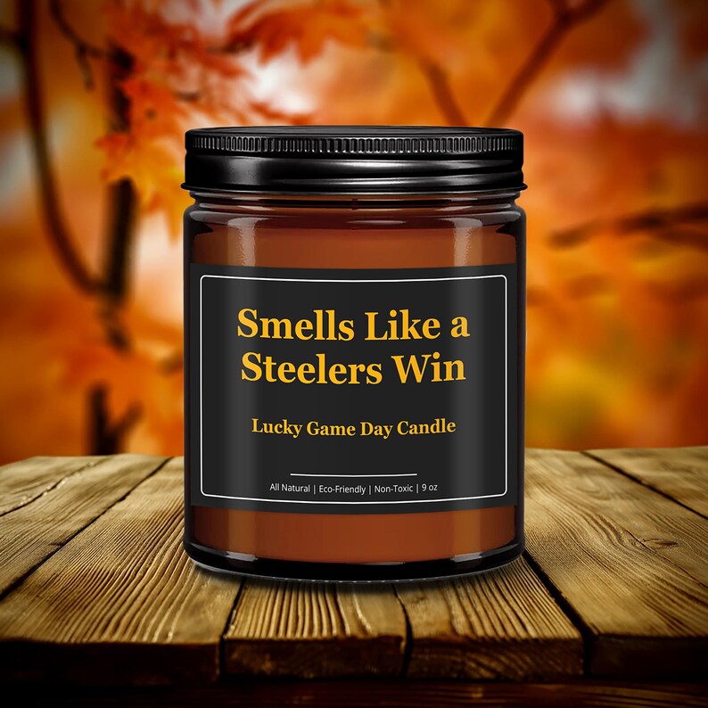 Wonderscape Wicks candle shop on Etsy presenting black lid 9oz amber jar soy wax candle with text smells like a Steelers win lucky game day candle on a wood table in a garden. Pittsburgh Football NFL sports gift for husband wife fiance boyfriend
